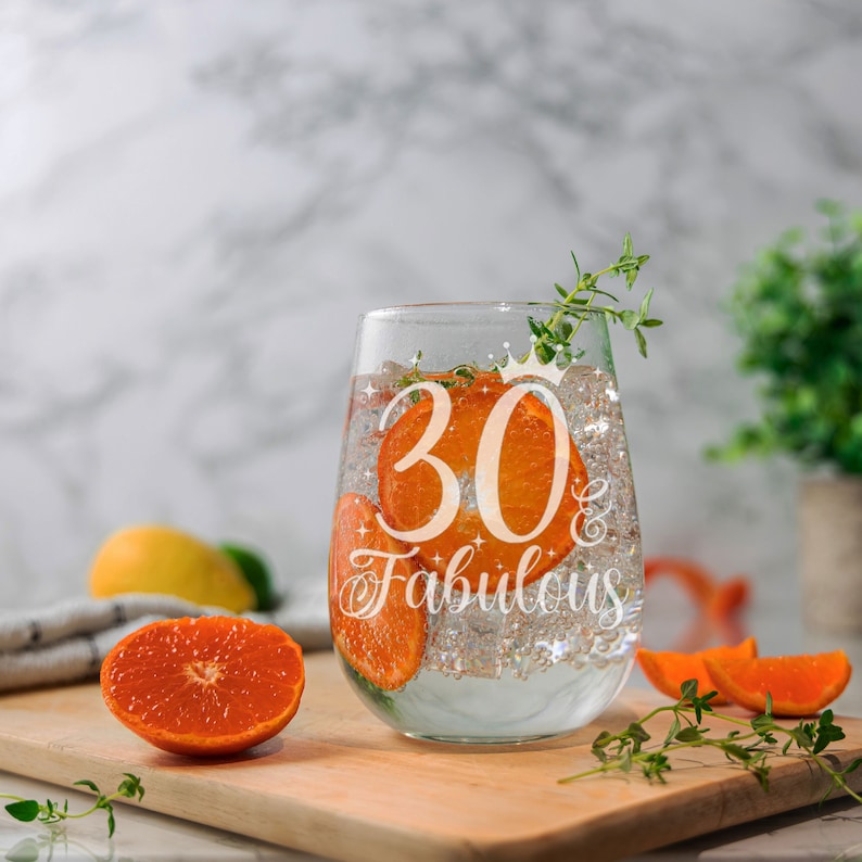 May include: A clear glass wine glass with the text "30 & Fabulous" etched on the side. The glass is filled with water and orange slices.
