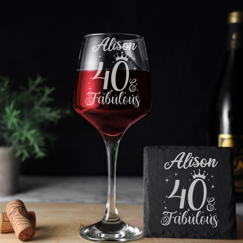 May include: A personalised wine glass with the text "Alison 40 & Fabulous" etched on it. The glass is filled with red wine and is sitting on a wooden surface. There is a black slate coaster with the same text on it.