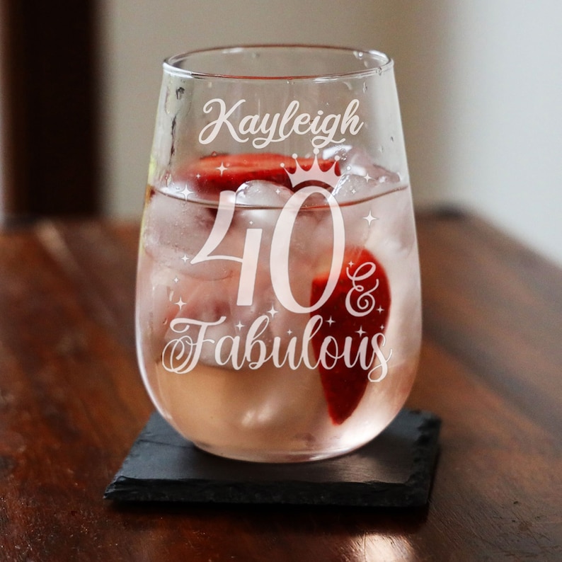 May include: A clear glass wine glass with the text "Kayleigh 40 & Fabulous" etched on the side. The glass is filled with a pink drink and ice.