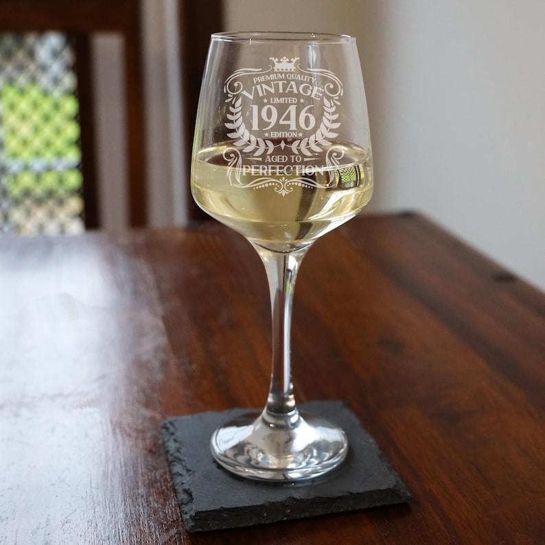 May include: A clear glass wine glass filled with a light-colored liquid, featuring an etched design that reads Premium Quality Vintage Limited 1946 Edition Aged to Perfection, complete with a crown and laurel wreath graphic on the front surface.