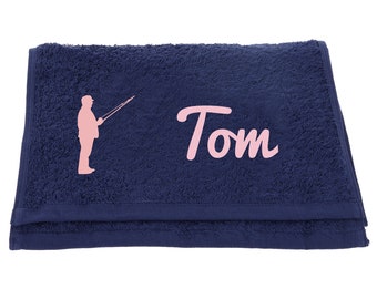 May include: A navy blue towel with a pink silhouette of a person fishing and the name "Tom" embroidered in pink.