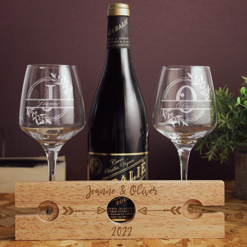 May include: A wooden wine bottle holder with two wine glasses. The holder has the names "Joanne & Oliver" and the year "2022" engraved on it. The glasses are engraved with the initials "J" and "O" and the names "Jeanne" and "Oliver".