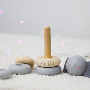 May include: A wooden stacking toy with grey and natural wood rings. The top ring has the name "Alice" written on it. The toy also includes a grey elephant with a smiling face.