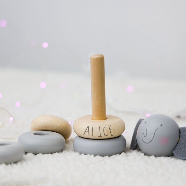 May include: A wooden stacking toy with grey and natural wood rings. The top ring has the name "Alice" written on it. The toy also includes a grey elephant with a smiling face.