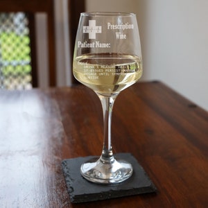 May include: A clear glass wine glass with a white inscription that reads "Prescription Wine" and "Patient Name:" with a red cross and a heart beat symbol. The glass is filled with white wine and is sitting on a black slate coaster.