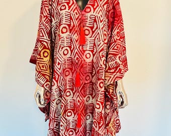 May include: A red and white patterned caftan dress with a matching head wrap. The dress features a V-neck, long sleeves, and a geometric design. Red tassels hang from the neckline. The outfit is displayed on a mannequin.