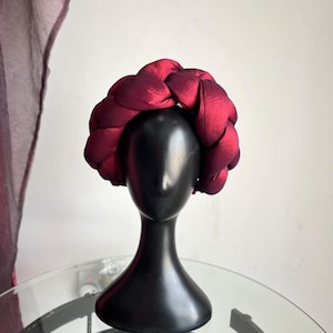以下が含まれることがあります： 黒いマネキンの頭の上に、ブルゴーニュ色の編み込みヘアバンドが置かれています。ヘアバンドは光沢のあるシルクのような素材で作られています。マネキンは黒い金属フレームのガラスのテーブルの上にあります。背景には、シアーなブルゴーニュ色のカーテンがあります。