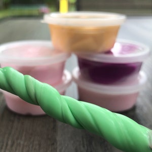 May include: Four small plastic containers filled with different colored slime. The containers are stacked on top of each other. A green slime is in the foreground.