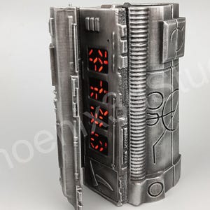 Könnte beinhalten: Ein silberfarbener, zylindrischer Requisite mit einer Scharniertür und einer Reihe roter digitaler Anzeigen. Der Requisite hat eine strukturierte Oberfläche und komplizierte Details.