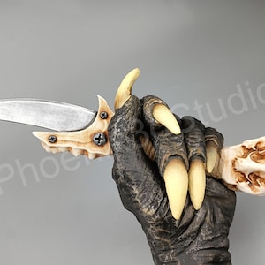 May include: A bone-like knife with a silver blade and a bone-like handle. The knife is held in a hand with claws.