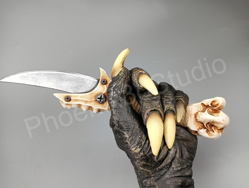 May include: A bone-like knife with a silver blade and a bone-like handle. The knife is held in a hand with claws.