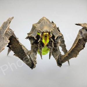 Può includere: Una figura di mostro articolata in plastica, dettagliata, di colore marrone e nero, con occhi verdi luminosi e una bocca spalancata. La figura ha un corpo segmentato e più arti.