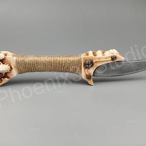 May include: A bone-handled knife with a brown twine wrap and a silver blade. The handle is sculpted to resemble a hand with a textured surface.