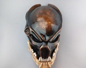 Può includere: Una maschera Predator nera e bronzo con un design dettagliato e una serie di denti affilati. La maschera ha una finitura bronzo e una visiera nera.