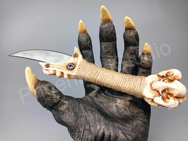 May include: A white and brown bone-like knife with a curved blade and a rope-wrapped handle. The knife is held in a hand with long, sharp claws.