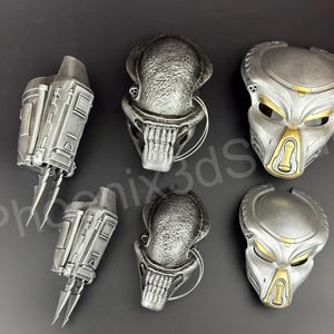 Puede incluir: Colección de artículos temáticos de Predator, que incluyen máscaras y cuchillas de muñeca, en plata y oro metálicos. Las máscaras tienen detalles intrincados y las cuchillas de muñeca tienen bordes afilados. Los artículos están dispuestos sobre una superficie gris oscuro.