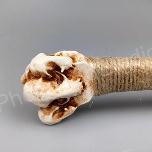 May include: A white ceramic sculpture with a rough, textured surface. The sculpture is shaped like a hand with brown rope wrapped around the wrist.