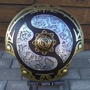 May include: A large, round, decorative shield with a brown, gold, and silver design. The shield has a central circular design with a stylized "S" in the center. The shield is on a stand with the text "DOTA 2" on it.