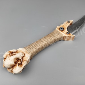 May include: A hand-crafted dagger with a bone handle, wrapped in twine, and a curved blade. The blade is made of metal and has a dark grey finish.