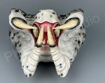 May include: A detailed, textured mask in shades of white, gray, and black, featuring large, curved ivory-colored tusks and a red interior. The mask has a reptilian or alien aesthetic, with a wide open mouth.