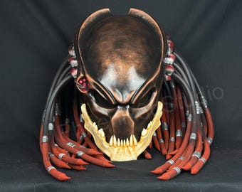 以下が含まれることがあります： ダークブロンズ色のヘルメットで、生物のようなデザインが特徴です。突き出た顎のライン、鋭いオフホワイトの歯、そして銀色の金属リングで装飾された、黒と赤の長く柔軟なドレッドヘアのような付属物が多数付いています。