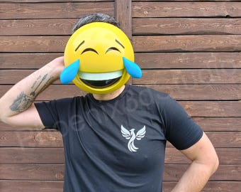 May include: A person wearing a large yellow emoji mask with blue tears and a wide smile. The person is wearing a black t-shirt with a white phoenix logo. The background is a wooden wall.