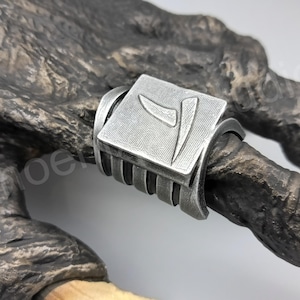 May include: A silver ring with a square design featuring a stylized symbol. The ring is worn on a finger with a claw-like appearance.