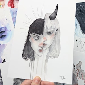 May include: A watercolor illustration of a split-faced figure, one side black-haired and the other white-haired with a horn. The artwork features a halo and dripping paint effect. Other art pieces are visible in the background.