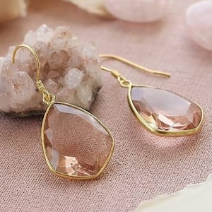 May include: A pair of teardrop-shaped morganite earrings set in 14k gold, featuring faceted stones with a soft pink hue. A small card with the text Genuine Morganite & 14k Gold is visible, alongside a cluster of raw crystals and a wax seal stamp.