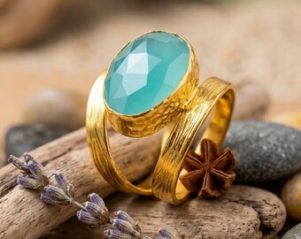 May include: A gold ring featuring a large, oval-shaped, faceted light blue gemstone set in a textured, hammered metal band. The ring has a split-shank design with two distinct bands that meet at the base of the central stone setting.