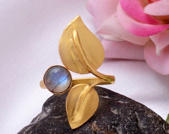 Può includere: Anello in oro con una pietra preziosa blu e due disegni a forma di foglia. L'anello è su una roccia grigio scuro.