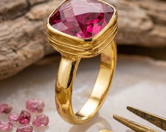 May include: A gold ring featuring a large, cushion-cut, deep pink gemstone set in a polished gold band. The ring is displayed alongside small, loose pink gemstone fragments, a piece of gold leaf, and metal tweezers on a textured surface.