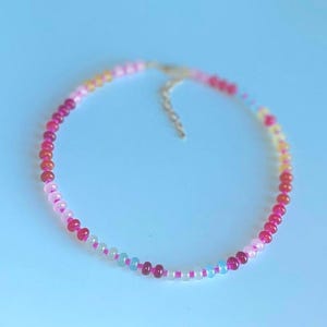 May include: A delicate necklace featuring a strand of colorful beads in shades of pink, red, yellow, and blue. The beads are strung together with a silver-colored chain and clasp. The necklace is displayed on a light blue background.