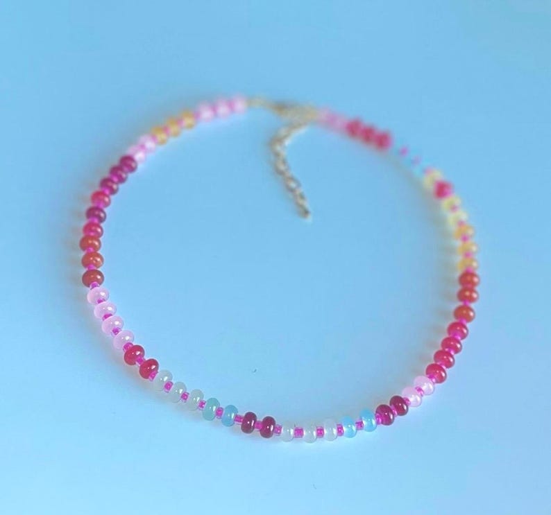 May include: A delicate necklace featuring a strand of colorful beads in shades of pink, red, yellow, and blue. The beads are strung together with a silver-colored chain and clasp. The necklace is displayed on a light blue background.