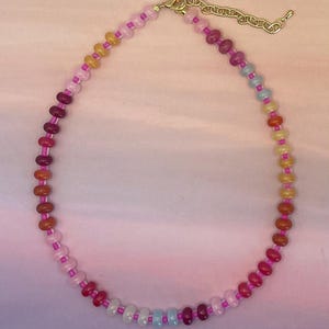 May include: A colorful beaded necklace with a gold-toned clasp and chain. The necklace features a gradient of colors, including pink, orange, yellow, blue, and purple beads. The beads are strung on a pink cord.