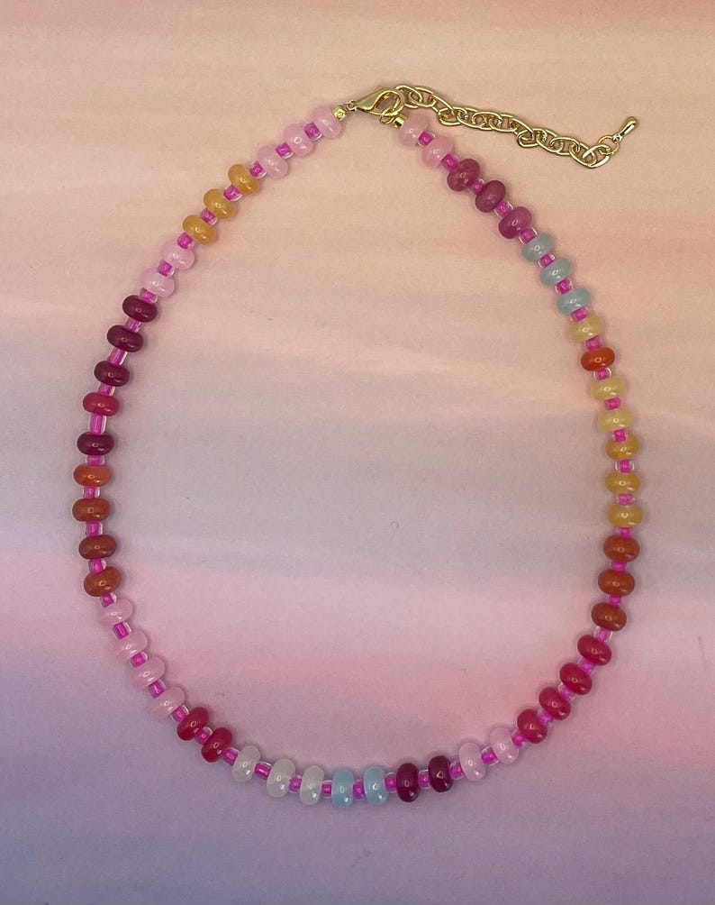 May include: A colorful beaded necklace with a gold-toned clasp and chain. The necklace features a gradient of colors, including pink, orange, yellow, blue, and purple beads. The beads are strung on a pink cord.