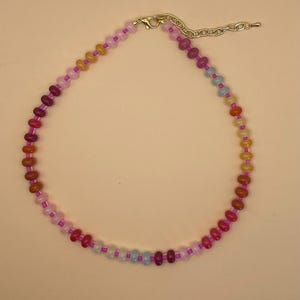 May include: A colorful beaded necklace with a variety of small, round beads in shades of red, orange, yellow, pink, and blue. The necklace has a gold-colored clasp and a chain for adjustable sizing.