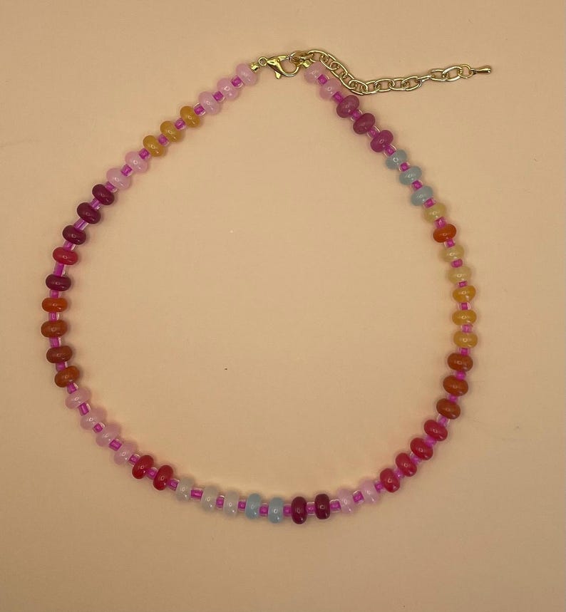 May include: A colorful beaded necklace with a variety of small, round beads in shades of red, orange, yellow, pink, and blue. The necklace has a gold-colored clasp and a chain for adjustable sizing.
