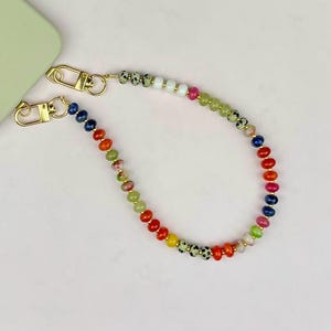 May include: A colorful beaded phone strap with gold-colored clasps. The beads are a mix of colors, including red, orange, yellow, green, blue, and pink. The strap is attached to a light green phone case.