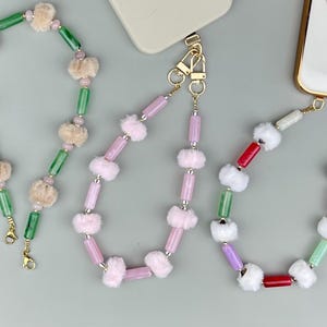 May include: A collection of colorful phone charms. The charms feature a variety of beads in green, pink, red, and white, interspersed with fluffy, cream-colored accents. Each charm has a gold-colored clasp for attachment.