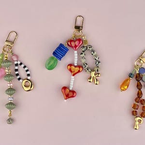 May include: Three beaded keychains with gold-tone clasps. One features a pink heart, green beads, and a black and white beaded loop. Another has red heart beads, a blue cylinder, and a dalmatian-spotted bead strand. The third has amber and brown beads.