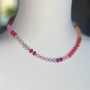 May include: A beaded necklace with a gradient of colors, from deep red to light pink and white. The necklace is displayed on a white mannequin. The beads are small and round, strung on a pink cord.
