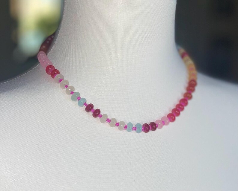 May include: A beaded necklace with a gradient of colors, from deep red to light pink and white. The necklace is displayed on a white mannequin. The beads are small and round, strung on a pink cord.