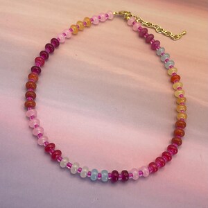 May include: A colorful beaded necklace featuring a variety of gemstone beads in shades of pink, orange, yellow, and blue. The necklace has a gold-colored chain and clasp. The beads are strung on a pink cord.