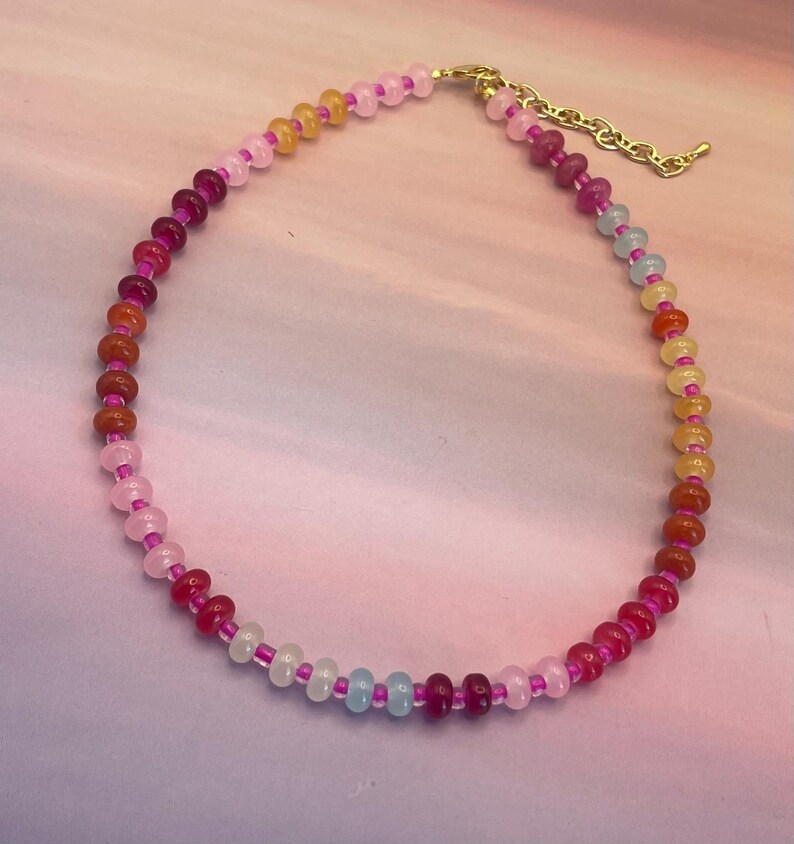 May include: A colorful beaded necklace featuring a variety of gemstone beads in shades of pink, orange, yellow, and blue. The necklace has a gold-colored chain and clasp. The beads are strung on a pink cord.