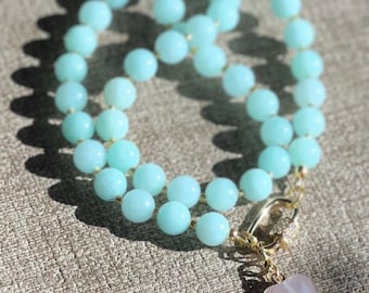 May include: A light blue beaded necklace with a gold clasp and a pink heart-shaped pendant. The necklace features round, light blue beads strung together. The pendant is a light pink heart with a gold border.