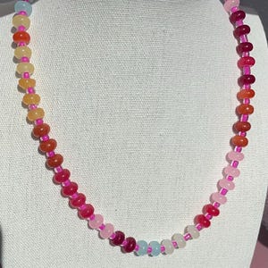 May include: A colorful beaded necklace with a gradient of colors. The necklace features small, round beads in shades of pink, red, orange, yellow, and blue, strung together with a pink cord. The necklace is displayed against a white background.