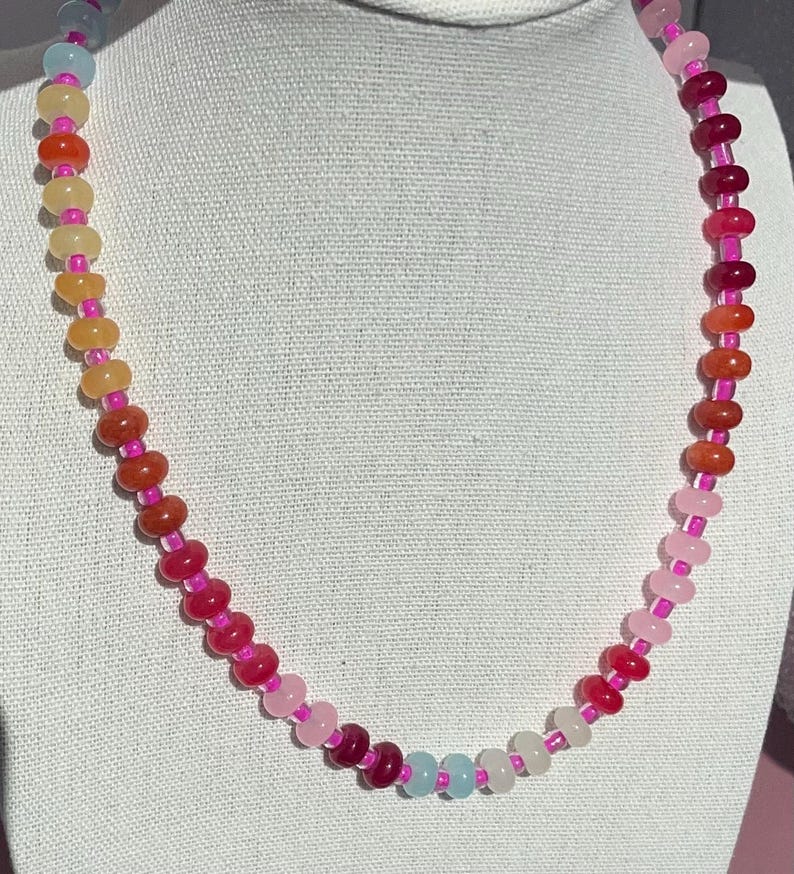 May include: A colorful beaded necklace with a gradient of colors. The necklace features small, round beads in shades of pink, red, orange, yellow, and blue, strung together with a pink cord. The necklace is displayed against a white background.