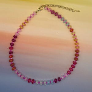 May include: A colorful beaded necklace with a gold-colored clasp and extender chain. The necklace features a variety of small, round beads in shades of pink, red, orange, yellow, and blue. The beads are strung on a pink cord.