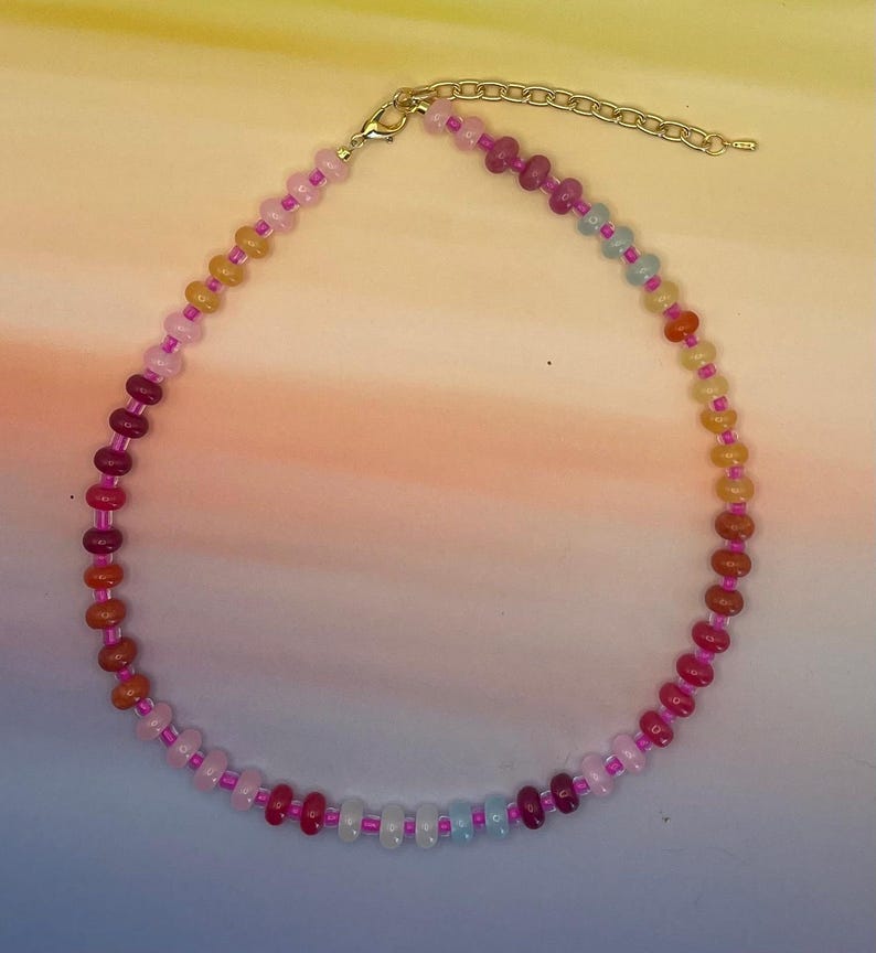 May include: A colorful beaded necklace with a gold-colored clasp and extender chain. The necklace features a variety of small, round beads in shades of pink, red, orange, yellow, and blue. The beads are strung on a pink cord.